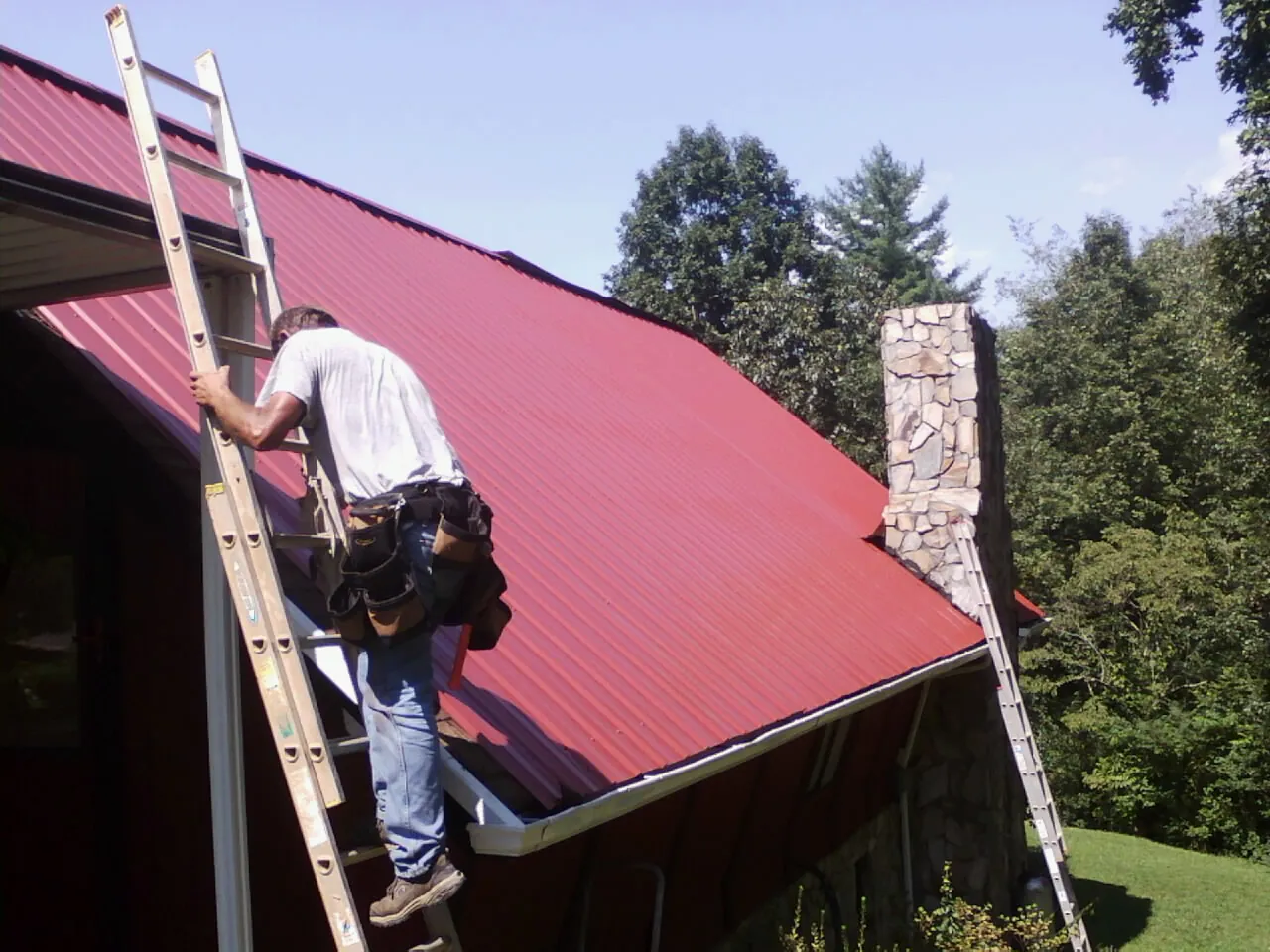 Roofer on a ladder beside a red metal roof and stone chimney — Soffit & Fascia Repair by Roofing Brothers in Huntingtown